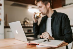 A young man works remotely in a kitchen, sipping coffee while his laptop and notebook are open on the table.
