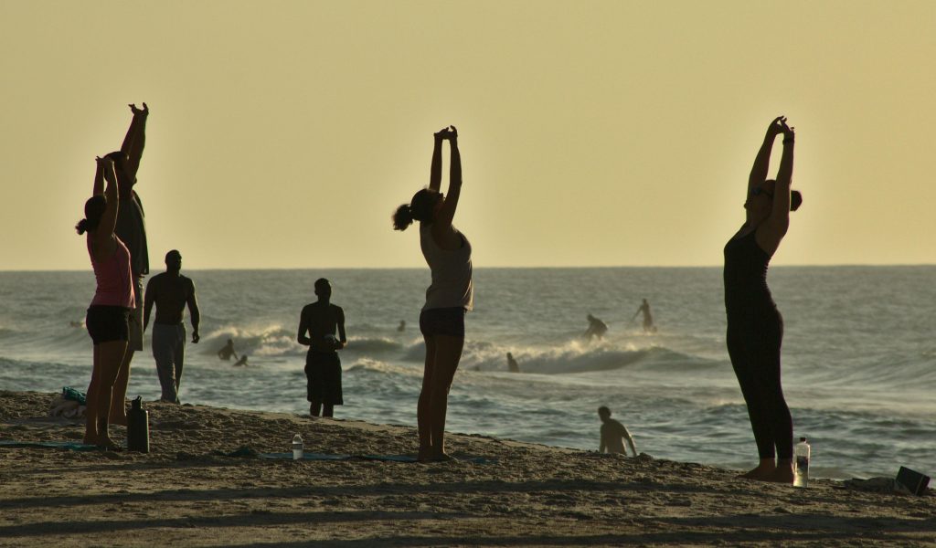 People practicing yoga on a North Carolina beach at sunset with surfers in the background.
