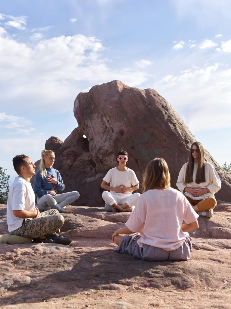 A group of adults meditating outdoors on a rocky landscape under a clear sky.